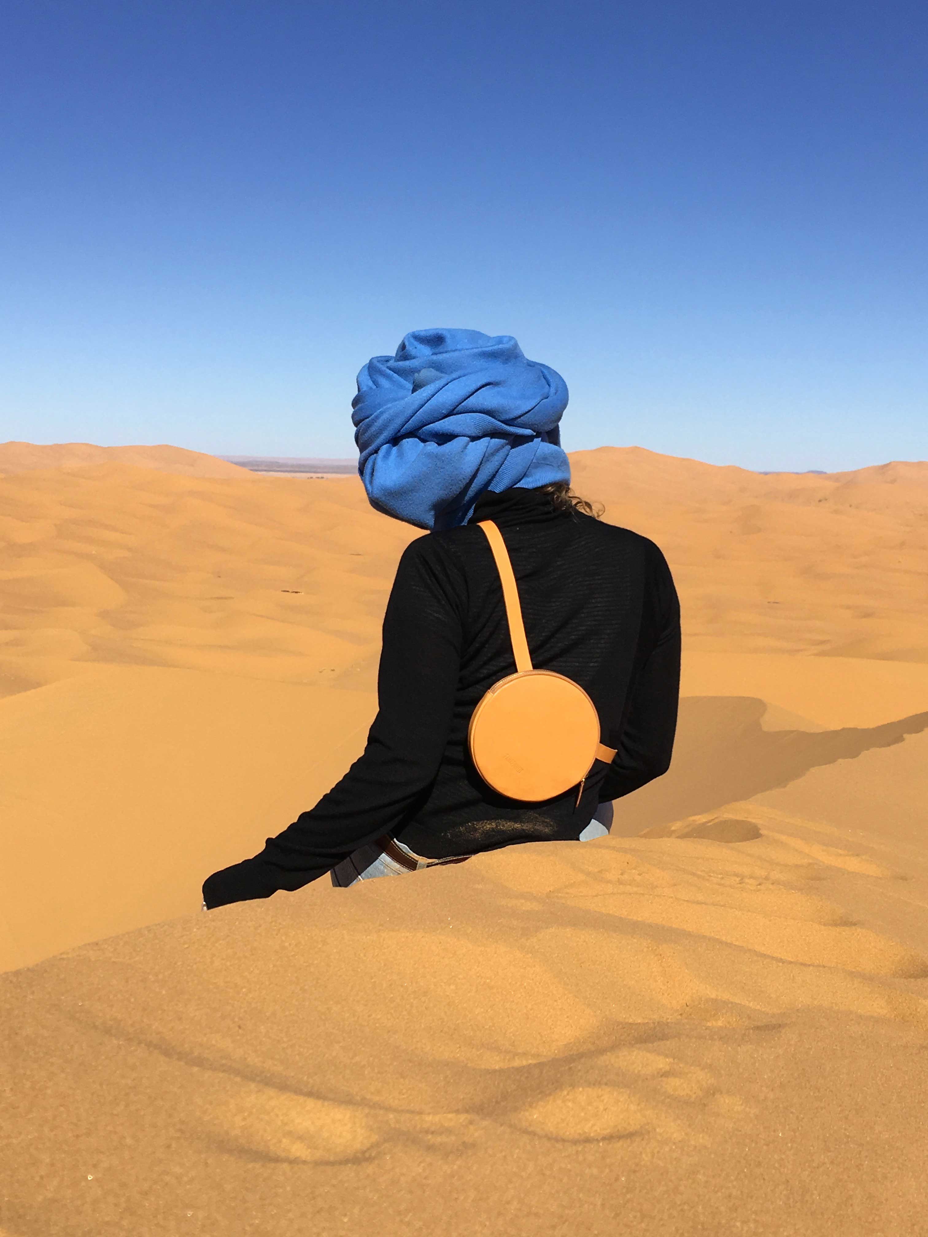 Woman in desert wearing a blue turban, a black turtleneck, a circle bag in sand color, with her back to the camera.