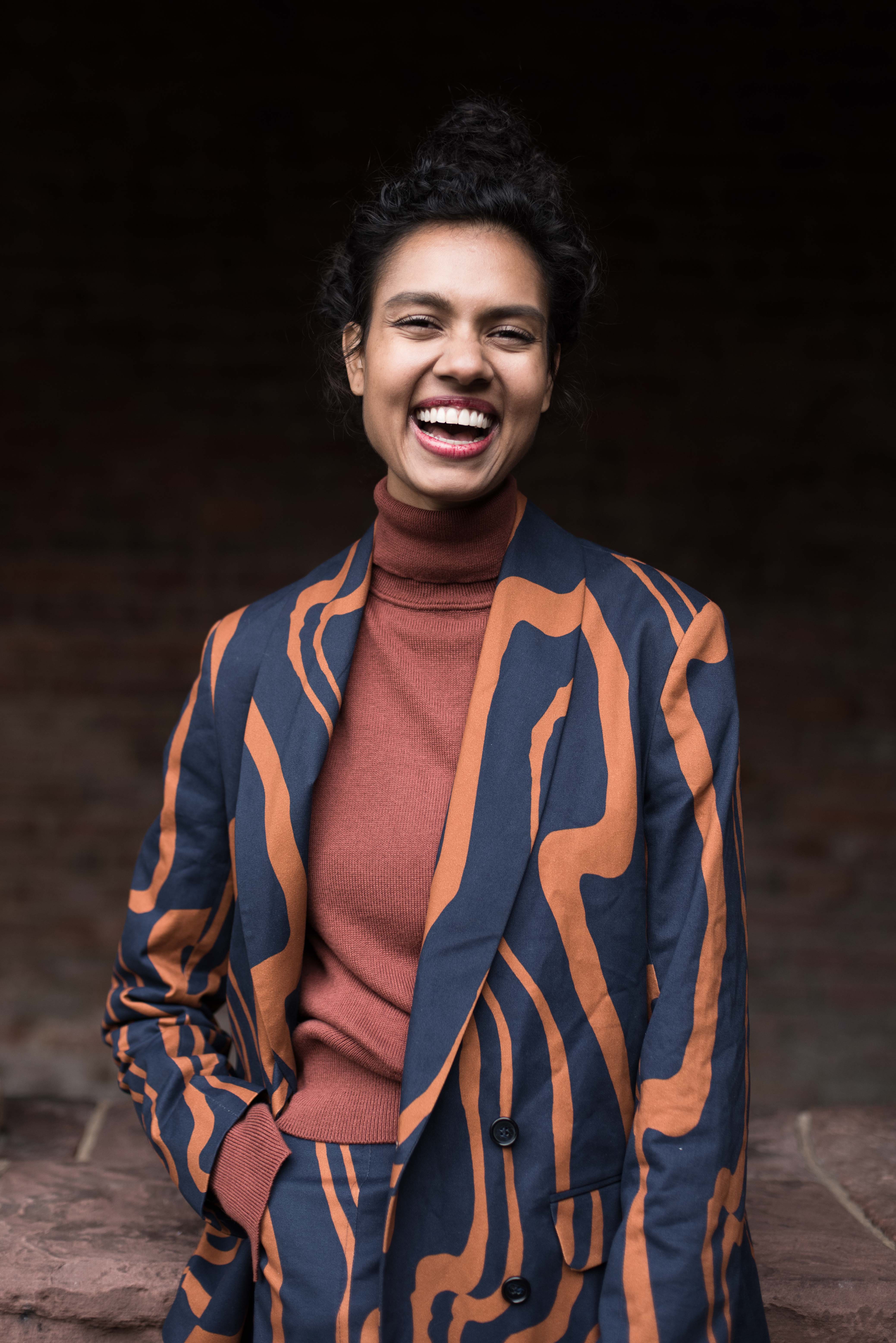 woman with hair pulled back, laughing. She is wearing a terracotta turtlneck and a navy and orange swirl print suit.